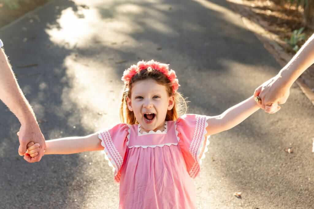 Little girl mid-swing laughing with arms outstretched holding her parents' hands in warm afternoon light.