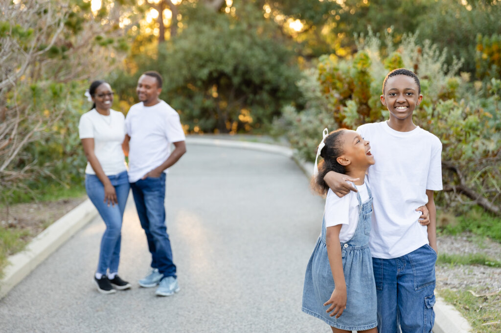 Brother and sister laughing together in the foreground with parents smiling in the background, outdoor family photos Perth